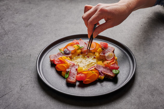 饮食女人的手用镊子把红辣椒粒放在一个深色盘子上 用自制的沙拉配上金枪鱼牛排 芒果酱 香草和香料 特写 食物装饰概念新鲜食物沙拉