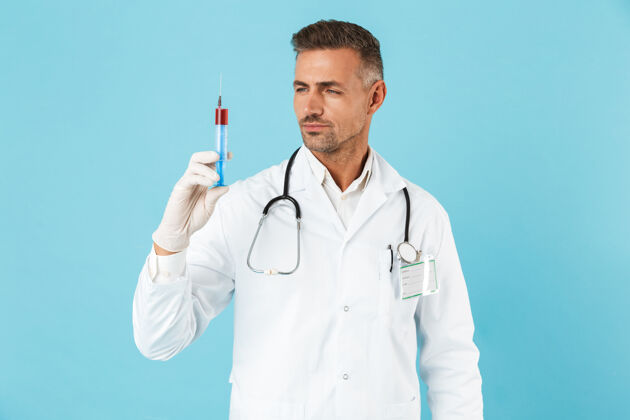 血液戴着听诊器戴着手套拿着注射器的欧洲医生的画像 孤立地站在蓝色的墙上专家医疗医院