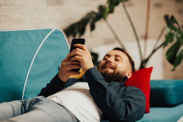 工作一个满脸笑容的大胡子男人一边用智能手机浏览有趣的视频 一边享受生活 一边躺在沙发上房间焦点在手上生活方式学习教育