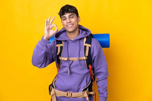 人一个年轻的登山者 背着一个大背包 站在隔离的黄色墙壁上 用手指画着“ok”的手势朋友手男人