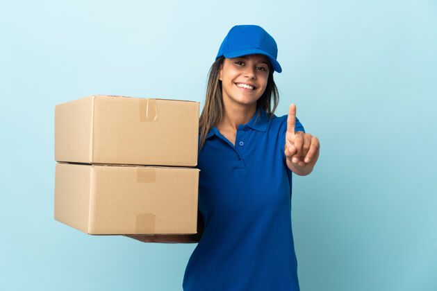 包装被隔离在蓝色墙上的年轻女送货员伸出手指拇指订单计数