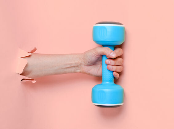 物体女的手拿着一个蓝色的塑料哑铃穿过撕破的粉色纸简约运动理念采取强壮手臂