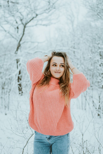 户外一个穿着粉色宽松毛衣和牛仔裤的漂亮女孩在寒冷的雪域森林里森林微笑年轻