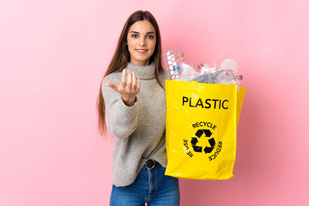 环境一位年轻的白人妇女拿着一个装满塑料瓶的袋子 在粉红色的背景下 邀请大家一起去回收手高兴你来了女人回收废物