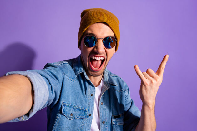 摇滚流行时尚开朗粗鲁尖叫的家伙自拍的自画像显示你有角的手指摇滚标志戴棕色帽子牛仔隔离紫色生动的色彩背景眼镜休闲符号
