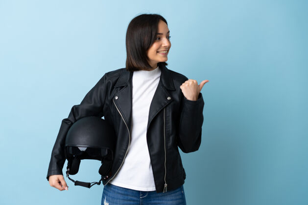 侧线女子手持摩托车头盔隔离在蓝色的墙上 指着侧面展示产品年轻皮革显示