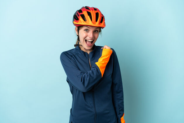 有趣一位年轻的骑自行车的女子被隔离在蓝色背景上庆祝胜利拳头欢呼力量