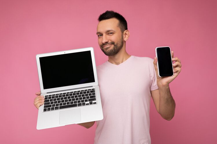 屏幕帅气的黑发男人拿着笔记本电脑和手机 在孤立的粉色背景下看着穿着t恤的相机电话在线键盘