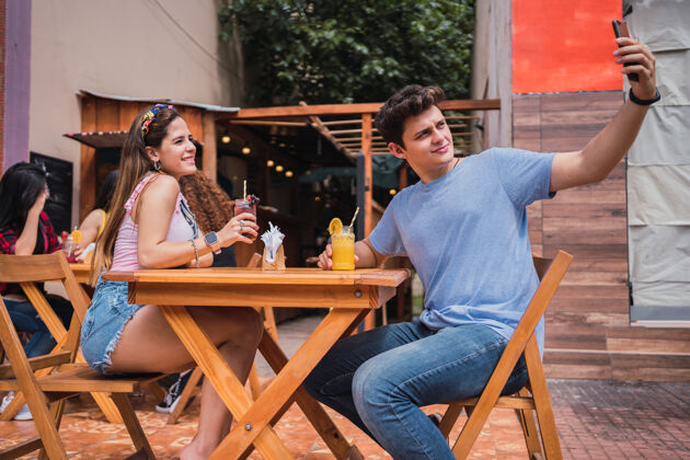 爱一对快乐的年轻夫妇拿着智能手机自拍——一对微笑的年轻夫妇坐在酒吧外面喝果汁浪漫约会男性