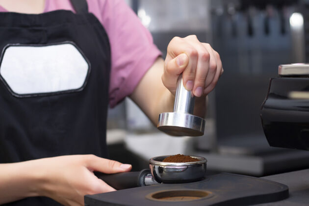制作特写照片中的女性手拿着一个金属捣固机和一个便携式过滤器在咖啡馆里喝咖啡咖啡师正在咖啡馆里准备压榨磨好的咖啡来冲泡浓缩咖啡或美式咖啡商店热的准备