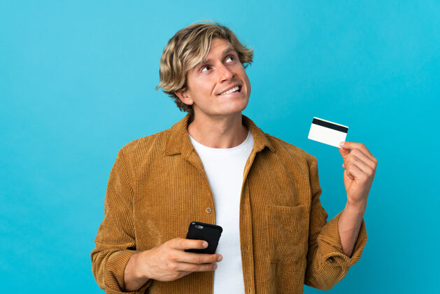 电话英国男人在孤立的蓝色背景下一边用手机购物一边用信用卡思考怀疑电话购买