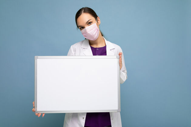 药品年轻迷人的护士 戴着防护面罩 穿着白色的医用外套 手里拿着一块隔离在蓝色背景上的空磁板白大衣女性白板