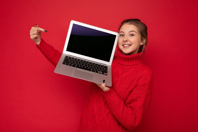 互联网照片中 一位美丽 满意 快乐的年轻女子手持笔记本电脑 看着摄像机 穿着红色毛衣 显示在显示器上 被隔离在红墙背景下笔记本电脑计算机技术