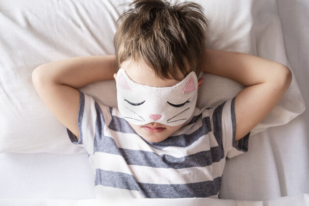 男孩高加索学龄前男孩穿着猫咪眼罩条纹睡衣躺在白色的床上可爱搞笑躺下
