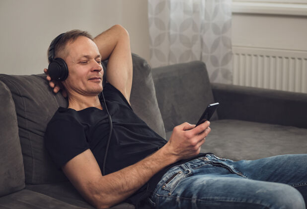 休息戴着耳机和智能手机的男人坐在沙发上时髦沙发男人