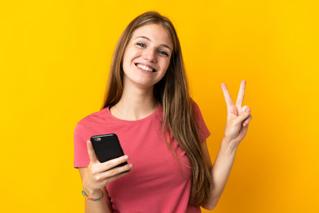 快乐年轻女子用手机孤立在黄色背景上微笑着 并显示胜利的迹象是的通讯交谈