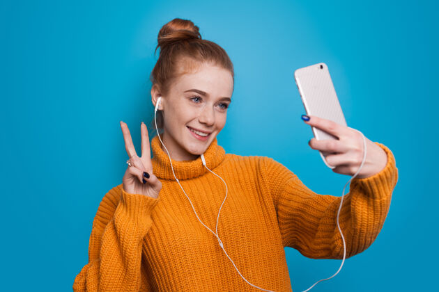 欢呼一位满脸雀斑 满头红发的年轻女子戴着耳机在蓝色工作室的墙上打电话问候别人毛衣模型技术