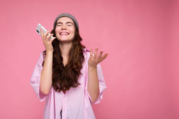 手机积极迷人的年轻黑发女子穿着时尚的粉红色衬衫和灰色帽子隔离在粉红色的背景下 手牵着手 用手机交流和录制语音信息粉色背景粉色衬衫黑发