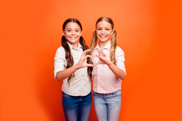 孩子正面开朗的肖像两个孩子女孩站在一起让心手指象征着亲情友好的家庭姐妹关系穿着休闲服装孤立的橙色背景头发年轻金发