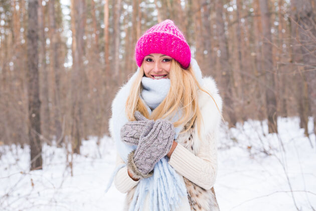 霜冻季节和人的概念-迷人的金发女人穿着白色外套和粉红色的帽子站在森林里快乐一月乐趣