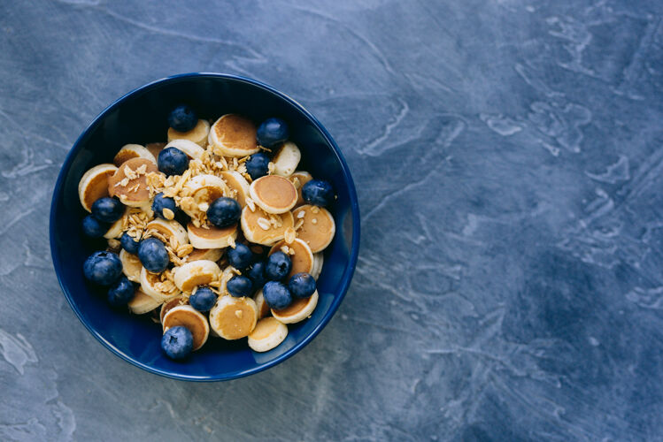 吃迷你煎饼麦片特写镜头 深蓝色碗里的迷你煎饼 枫叶糖浆蜂蜜和蓝莓面包房碗蓬松