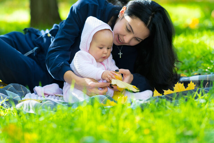 阳光可爱的小宝宝在公园里玩秋叶 妈妈在毯子上搂着她关系女孩家庭