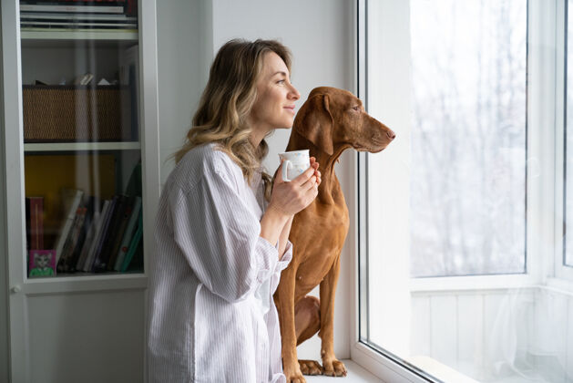 宠物快乐的女主人和狗坐在窗台上 端着一杯咖啡 望着窗外咖啡主人窗台