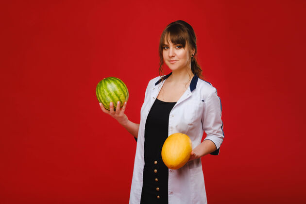 维生素一位身穿白大褂 手拿水果的女医生护士在红色背景上摆姿势 甜瓜 西瓜 饮食西瓜女孩