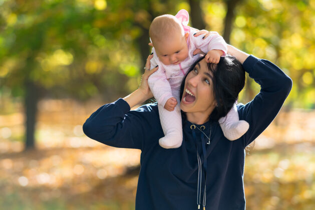 婴儿欢笑的母亲让她的小女儿骑在背上 因为他们一起走过一个多彩的秋天森林乐趣宝贝情感