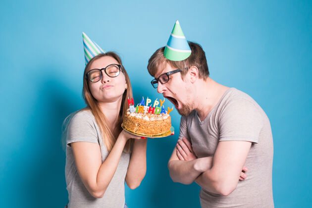 爱一对奇怪的年轻夫妇戴着纸帽子的女孩和男孩想咬下一块祝贺蛋糕疯狂节日庆祝