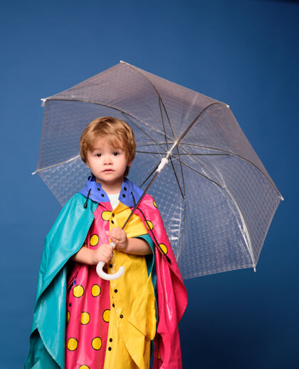 乐趣微笑的小男孩在玩树叶 看着照相机开朗的穿雨衣的男孩带着彩色雨伞玩具童年概念11月