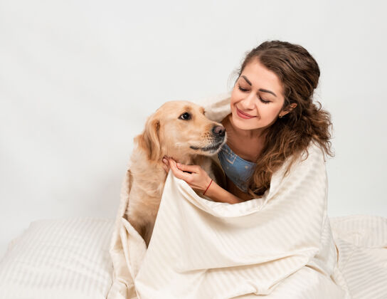 可爱女孩抱着她的拉布拉多猎犬 裹着一条白色的毯子欢呼床成人
