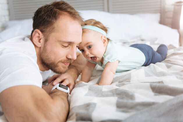 父母熟悉现代科技活泼可爱的小宝宝和爸爸躺在床上 看着智能手表 表达着兴趣和喜悦谎言世代孩子