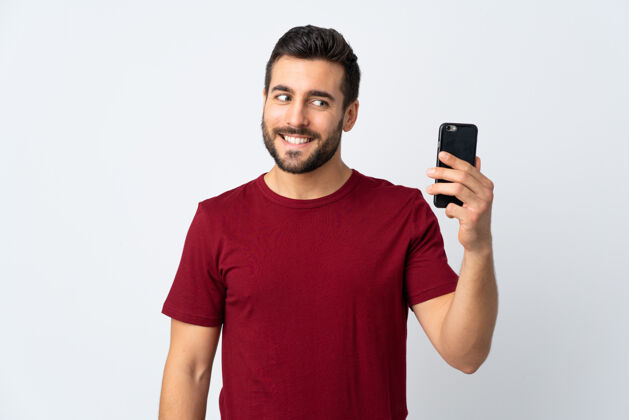 手机年轻帅哥留着胡子用手机隔离在白色上带着幸福的表情乐趣通讯表情