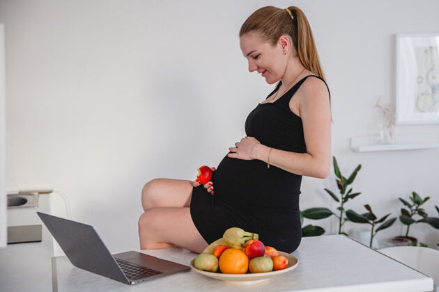 盘子在白色厨房里 一位身穿黑色连衣裙的年轻金发孕妇正坐在桌子上 吃着水果 看着一张桌子笔记本电脑高高质量的照片房间新鲜桌子