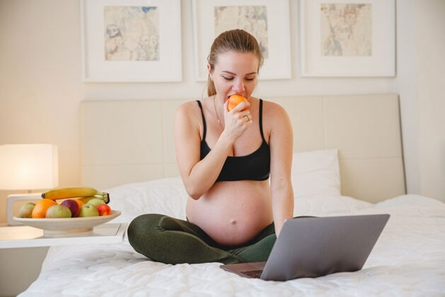 水果一个年轻的金发孕妇坐在床上 一边吃水果一边看着她笔记本电脑高高质量的照片冠状病毒期间吃