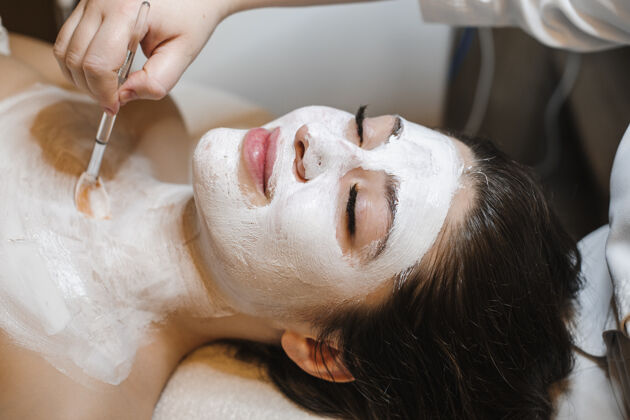 脸一个年轻迷人的女人闭着眼睛靠在一张有身体和白色面膜的水疗床上的特写镜头皮肤护理头发泥