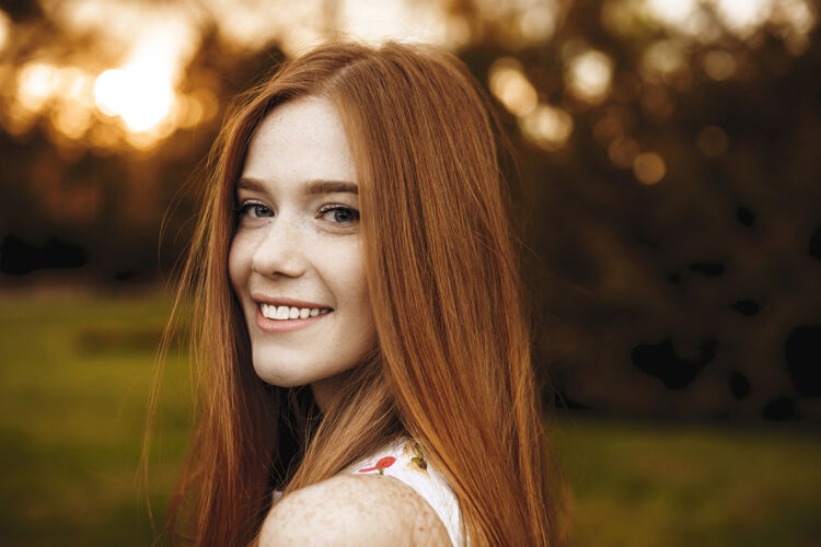 皮肤一个满脸雀斑的红发女孩在镜头前的特写照片 在外面夕阳的映衬下 她在肩上笑着女性户外特写