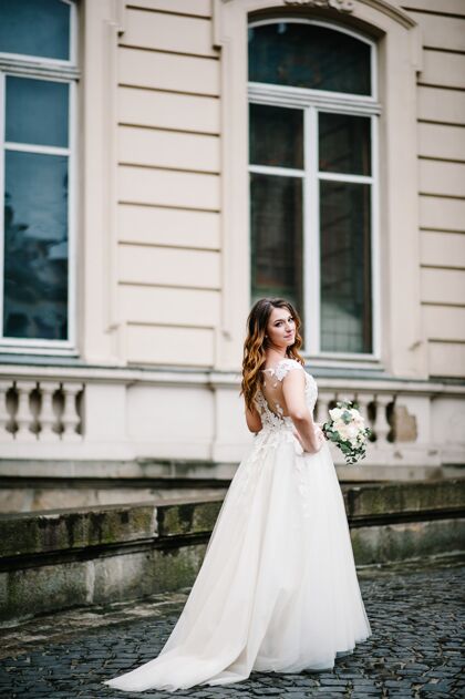 年轻身着婚纱的新娘肖像 带着一束鲜花 站在外面的旧建筑附近 复古宫殿在户外花城堡优雅