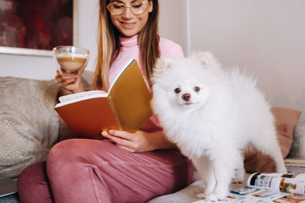 女人一个穿着睡衣的女孩在家里和她的狗斯皮策一起看书 狗和它的主人正在沙发上休息 看一本书书籍.家庭家务活阅读女性成人