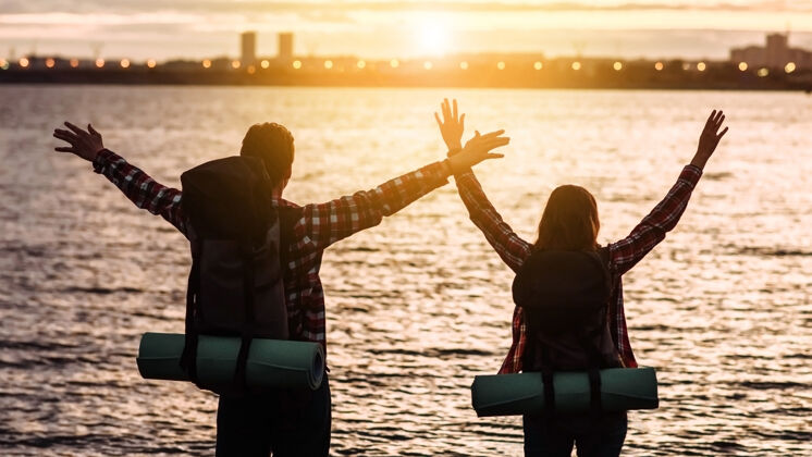 高两个背着背包的徒步旅行者 一男一女 在日落的海面上举手度假海男爱