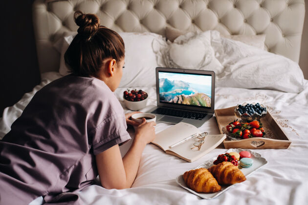 吃一个女孩晚上坐在床上 手里拿着智能手机 吃着草莓 一个女孩在床上睡觉前吃着糖果电话早餐工作