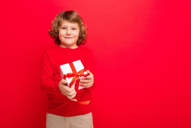 卷发照片拍摄的快乐微笑的金发卷发男孩孤立在红色背景墙穿着红色毛衣举行礼盒孩子祝贺快乐