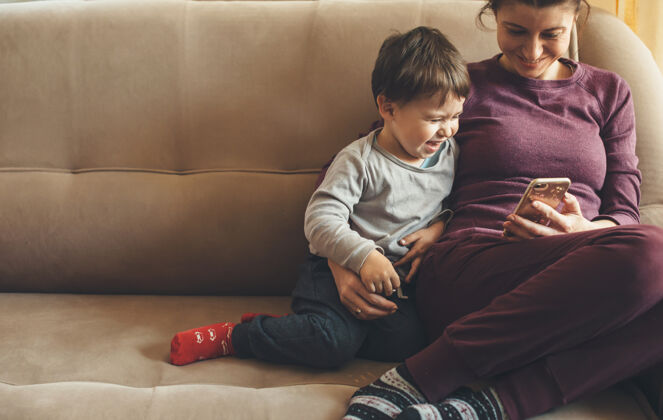 妈妈高加索妈妈和她的小男孩用手机坐在沙发上开心地笑着冠状病毒父母检疫