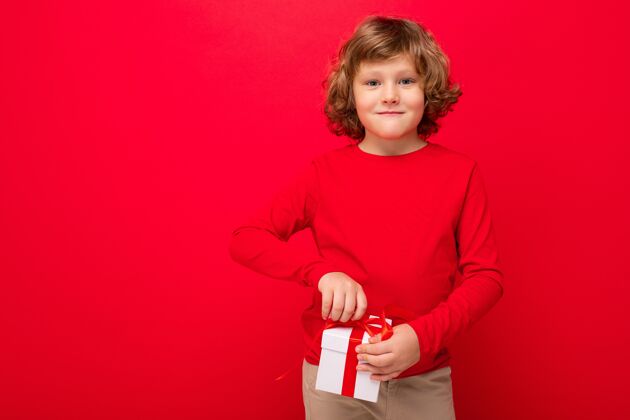 节日照片中的快乐微笑金发卷发的男孩孤立在红色背景墙上穿着红色毛衣拿着礼盒微笑礼品盒快乐