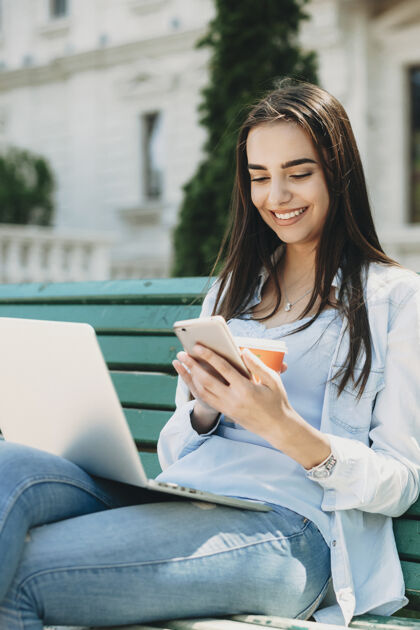 长凳一个年轻迷人的女学生坐在学校对面的长凳上 看着智能手机微笑 腿上拿着一台笔记本电脑休闲青少年独自