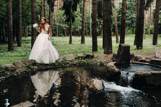 花束一位优雅的新娘身穿白色礼服 戴着手套 手持花束 站在森林的小溪边优雅手套婚礼