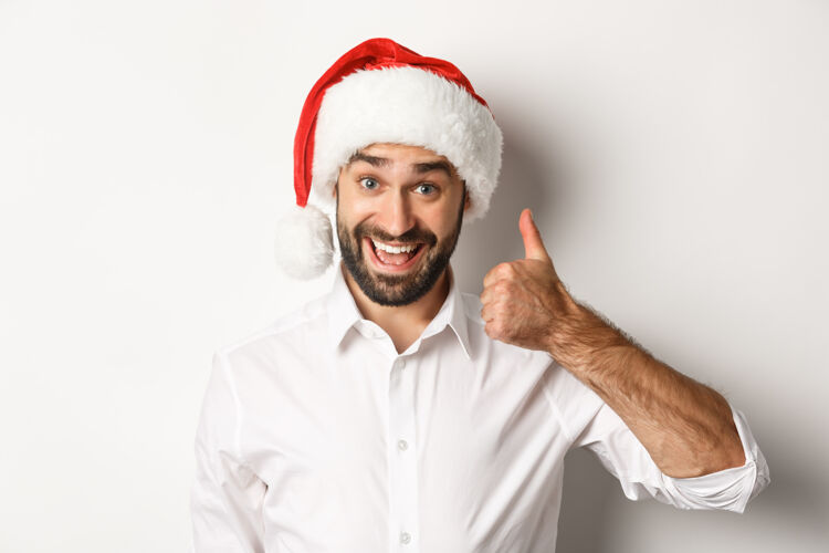 男人聚会 寒假和庆祝活动概念关闭-戴着圣诞帽的满脸胡须的男子竖起大拇指 表示满意 并喜欢一些好的白色背景节日圣诞老人时尚