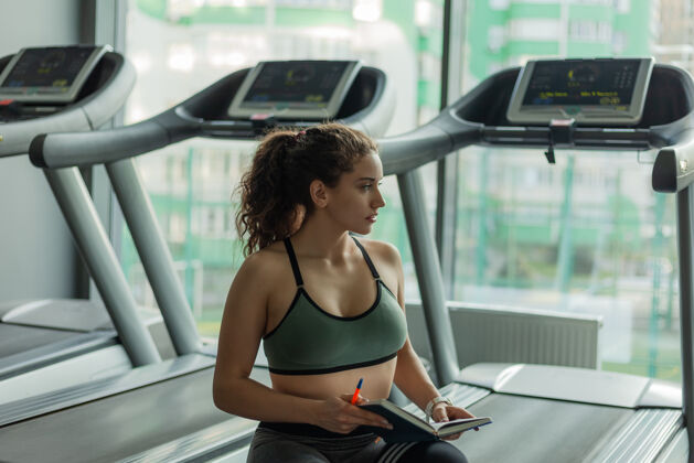 室内一位健康的年轻女子在一本培训书上写下了在体育训练中取得进一步进展的信息健身房健活方式概念你的锻炼计划健身女人运动体重