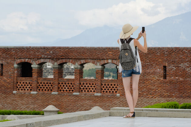 墙一个戴着帽子背着背包的女孩在拍一张老照片堡垒.view从后面摄影师天空背部